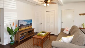 A living room with a television on a wooden stand, a sofa, a coffee table, and a ceiling fan. - Comfy Beach Studio for 2 - Only a short walk to the beach. (Cape Canaveral)