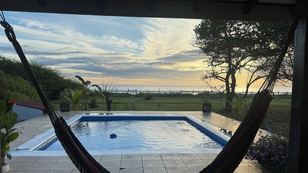 A hammock over a swimming pool with a view of the sea and a sunset.