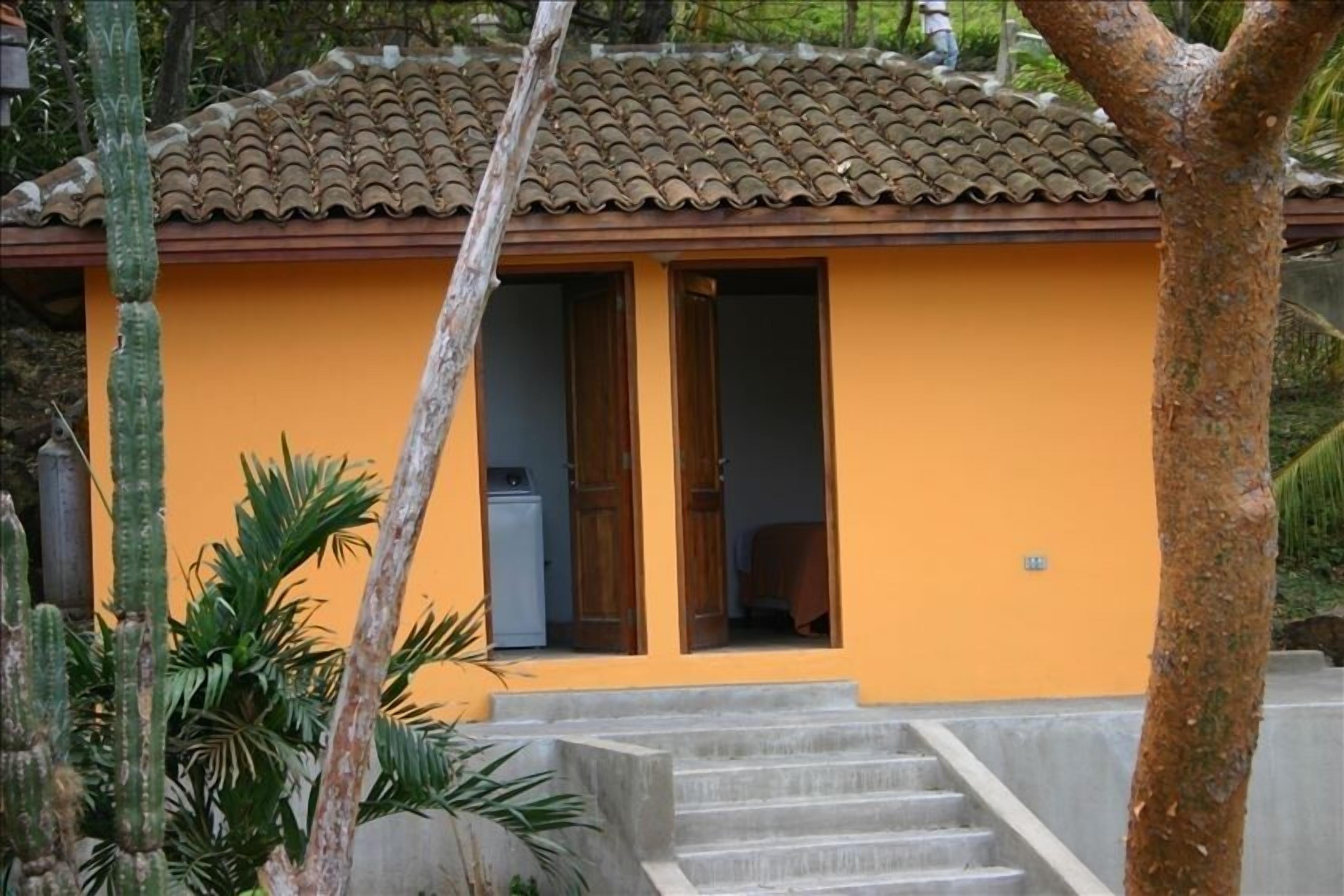 Un edificio pequeño con techo de tejas y exterior de color naranja brillante. Se observan dos puertas y una escalera de hormigón que conduce a la entrada.