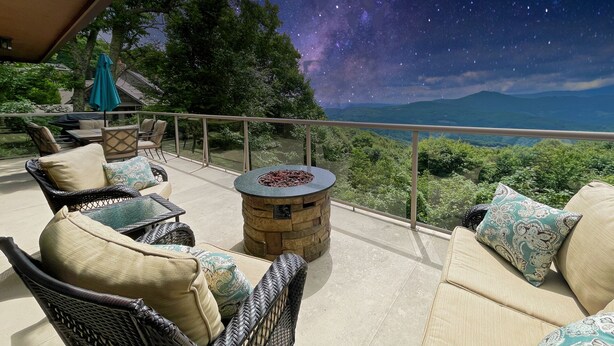 An outdoor seating area with wicker furniture and a stone fire pit, overlooking a mountainous landscape and a starry sky.
