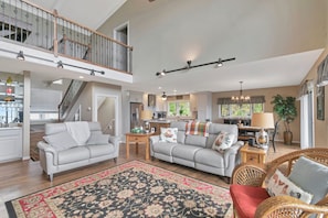 A spacious living area with a high ceiling, a staircase, and a kitchen in the background.
