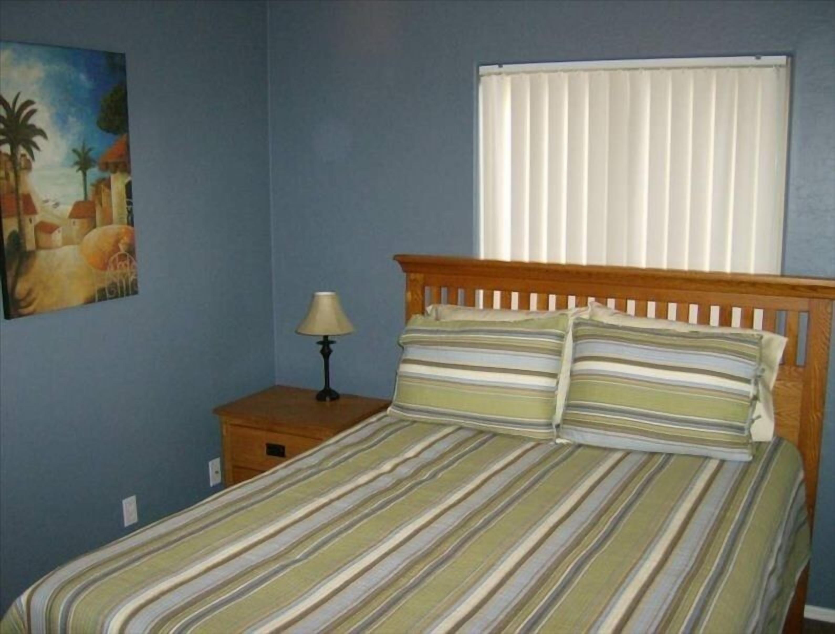 A bedroom with a bed, a nightstand, a lamp, a window with blinds, and a painting on the wall.