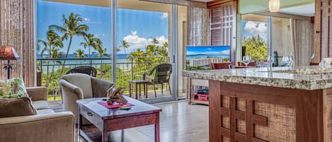 A spacious living area with a large sliding glass door leading to a balcony with a view of the ocean and palm trees. There is a ceiling fan, a coffee table with decorative items, and a bar area with a woven partition.