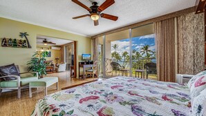 Una camera d'albergo con un letto grande, una ventola a soffitto, un balcone con vista su palme e sull'oceano, e un'area pranzo con una TV.