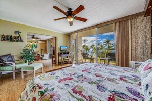 Hotelkamer met een groot bed, een plafondventilator, een balkon met uitzicht op palmbomen en de oceaan, en een zithoek met een televisie.