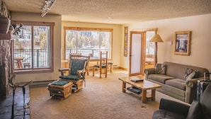 A cozy living room with a fireplace, a sofa, a dining table, and a view of the outdoors through large windows. - Spacious Luxury Condo Private Hot Tub and Sanua in Unit 1000' to ski lift (Keystone)