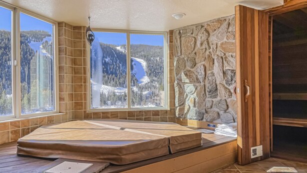 A room with a wooden floor, a large window with a view of mountains and a lake, and a stone wall. - Spacious Luxury Condo Private Hot Tub and Sanua in Unit 1000' to ski lift (Keystone)