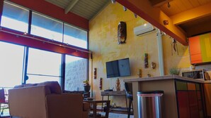 A spacious living area with a sofa, a TV mounted on the wall, and a kitchen area with cabinets and a microwave. - Artistic Top-Floor Tropical Penthouse – Steps to Maui’s Best Beach (Kihei)