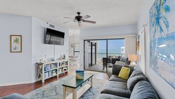 A living room with a sofa, a coffee table, a bookshelf, and a TV. There is a balcony with a view of the ocean.