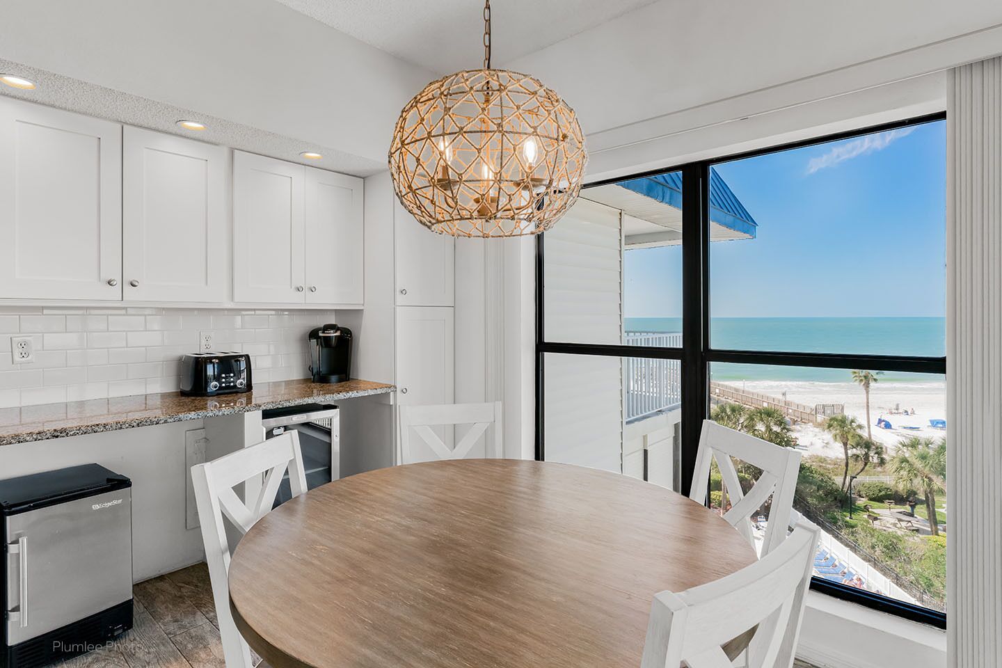 Una cocina moderna con armarios blancos, una mesa redonda de madera y un amplio ventanal con vistas a la playa.