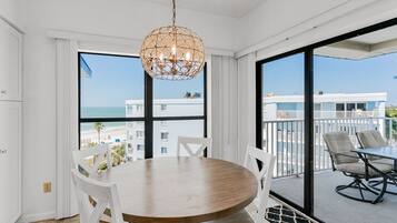 A dining area with a round table and chairs, large windows with a view of the beach and buildings, and a balcony with outdoor seating.