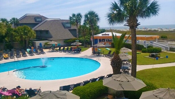 A pool area with lounge chairs and umbrellas, surrounded by palm trees and a building with a sloped roof. - Precious Island Cottage (Fripp Island)
