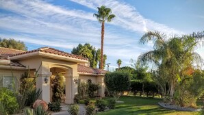 Una casa residencial con techo de tejas, césped cuidado y diversas plantas y árboles.
