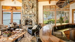A cozy living room with a stone fireplace, a wooden ceiling, and large windows overlooking a lake.