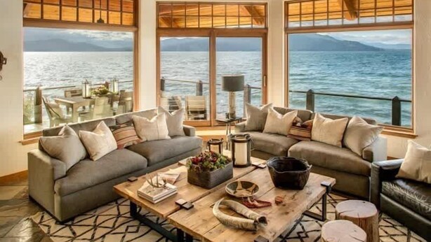 A spacious living area with a large window overlooking a body of water, a cozy sofa set, a wooden coffee table, and a patterned rug.
