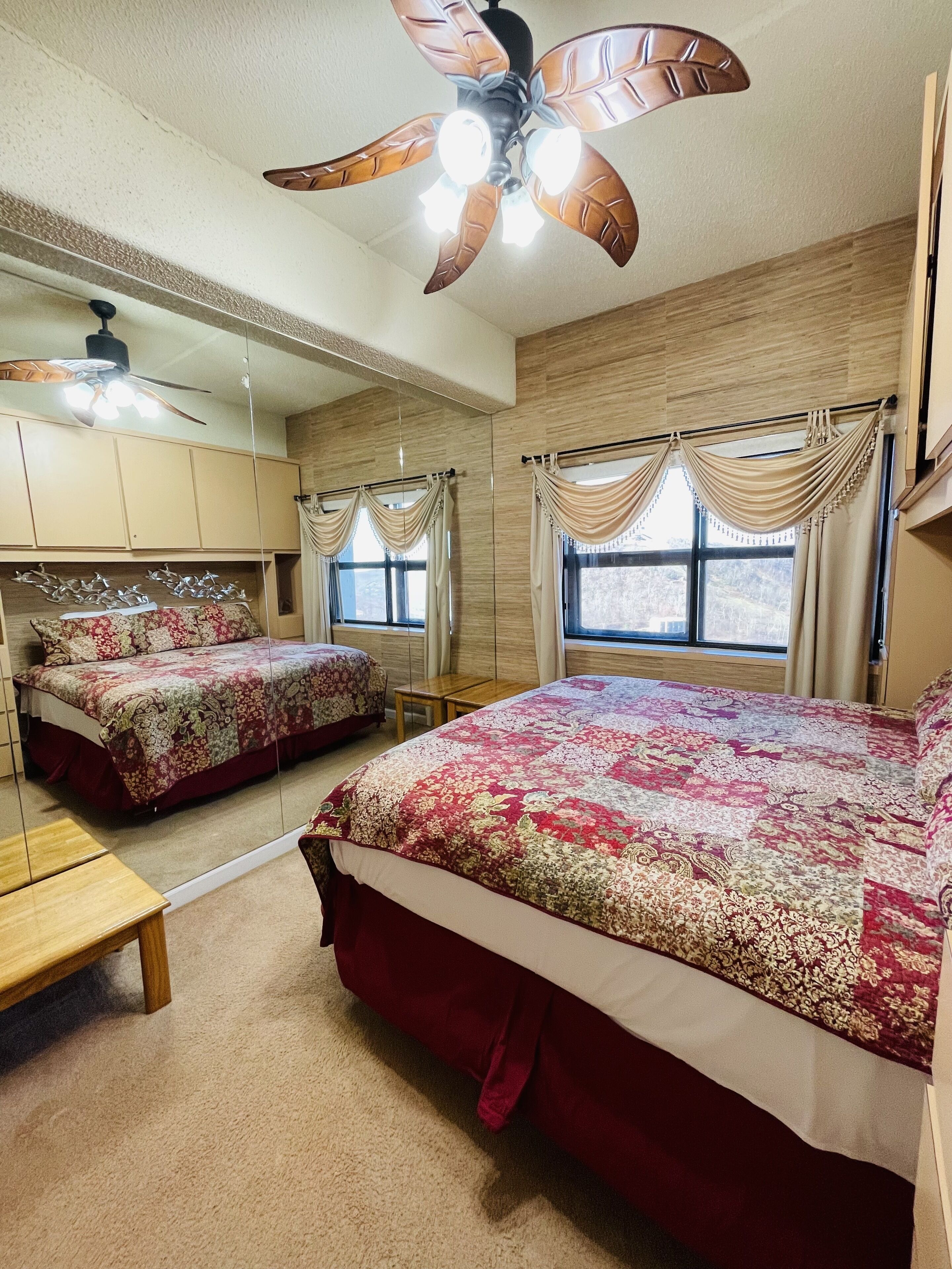 Une chambre à coucher avec un grand lit, un ventilateur de plafond, un miroir et une fenêtre avec des rideaux.