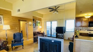 Ein geräumiges Wohnzimmer mit Kamin, Fernseher und Deckenventilator. Es gibt eine Küchenzeile mit Holzschränken und einer Mikrowelle.