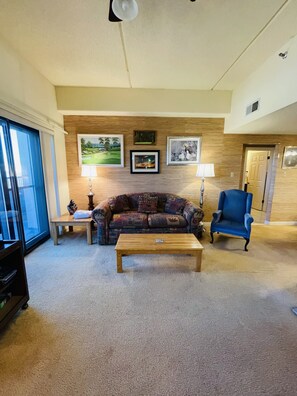 Uma sala de estar com uma parede de madeira, uma poltrona azul, um sofá estampado, uma mesa de centro e uma televisão.