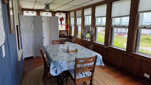 En matplats med ett bord dukat för två personer, trästolar, ett mönstrat bordsduks, en fläkt och stora fönster med persienner.