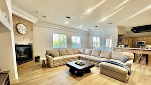Amplia sala de estar con un sofá seccional en forma de L, una mesa de centro y una chimenea. Amplias ventanas ofrecen luz natural y vistas al exterior.