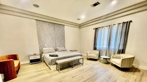 Un dormitorio moderno con una cama grande, mesitas de noche, un banco, dos sillones y una ventana con cortinas.