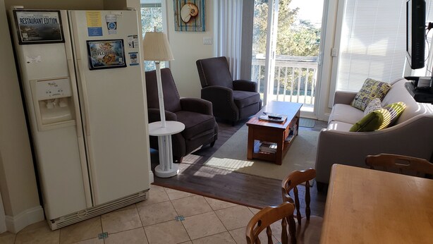 Ein Wohnzimmer mit einer Couch, Sesseln, einem Couchtisch und einem Esstisch. Ein Kühlschrank mit Magneten und ein Wasserspender sind ebenfalls vorhanden. Durch eine Schiebetür gelangt man in einen Außenbereich.