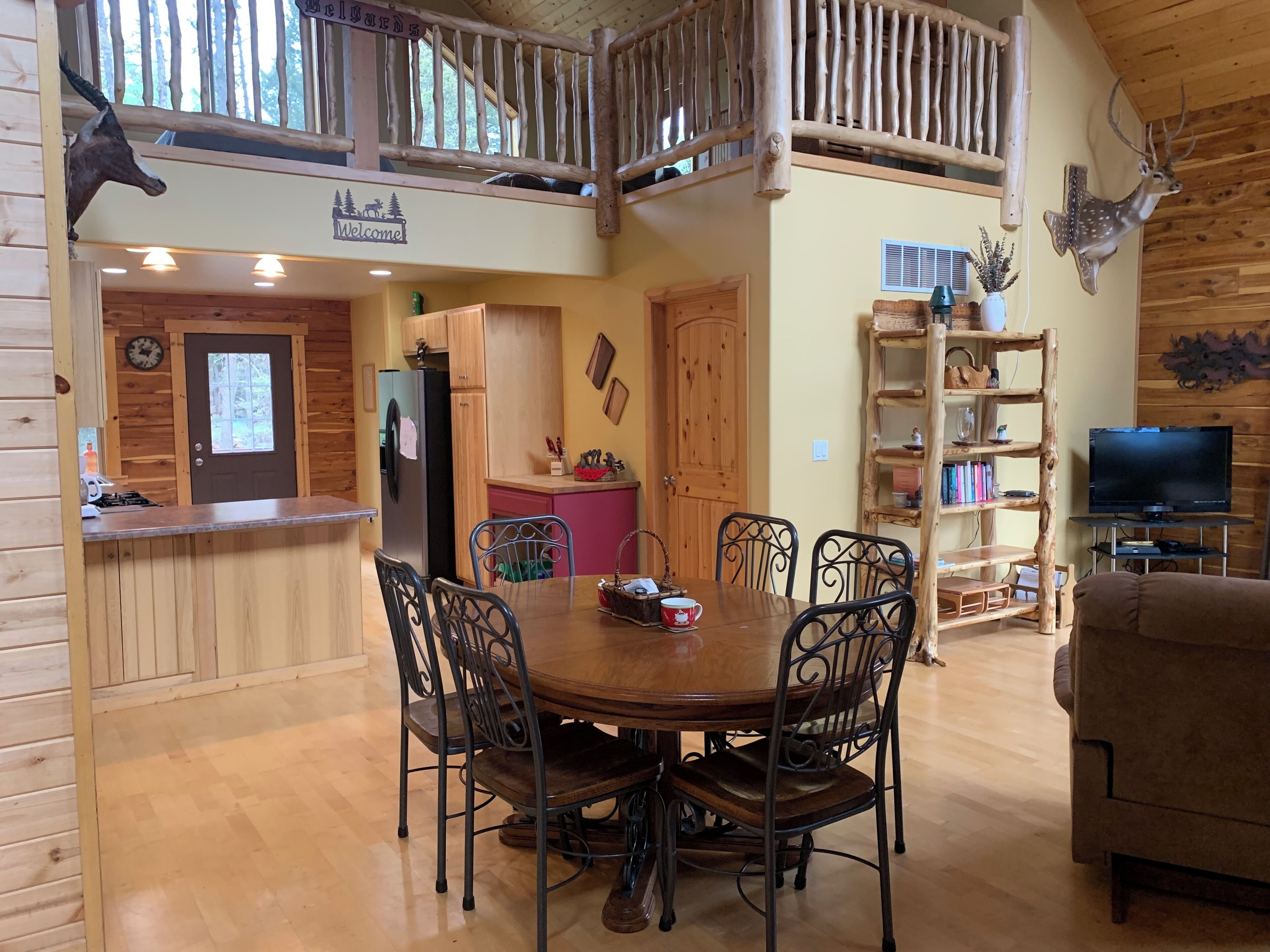 A wooden cabin interior with a dining area, kitchen, and a loft with a balcony.