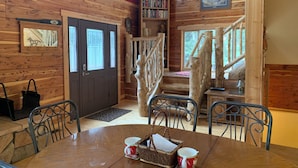 Interno di una baita in legno con un tavolo da pranzo, sedie, una scala, una libreria e una porta con una finestra.
