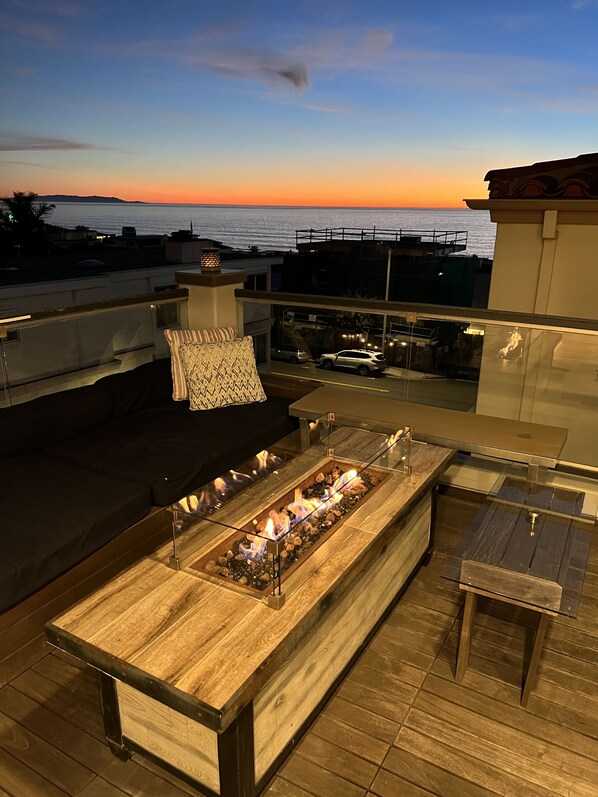 A rooftop terrace with a fire feature, seating area, and a view of the ocean at sunset.