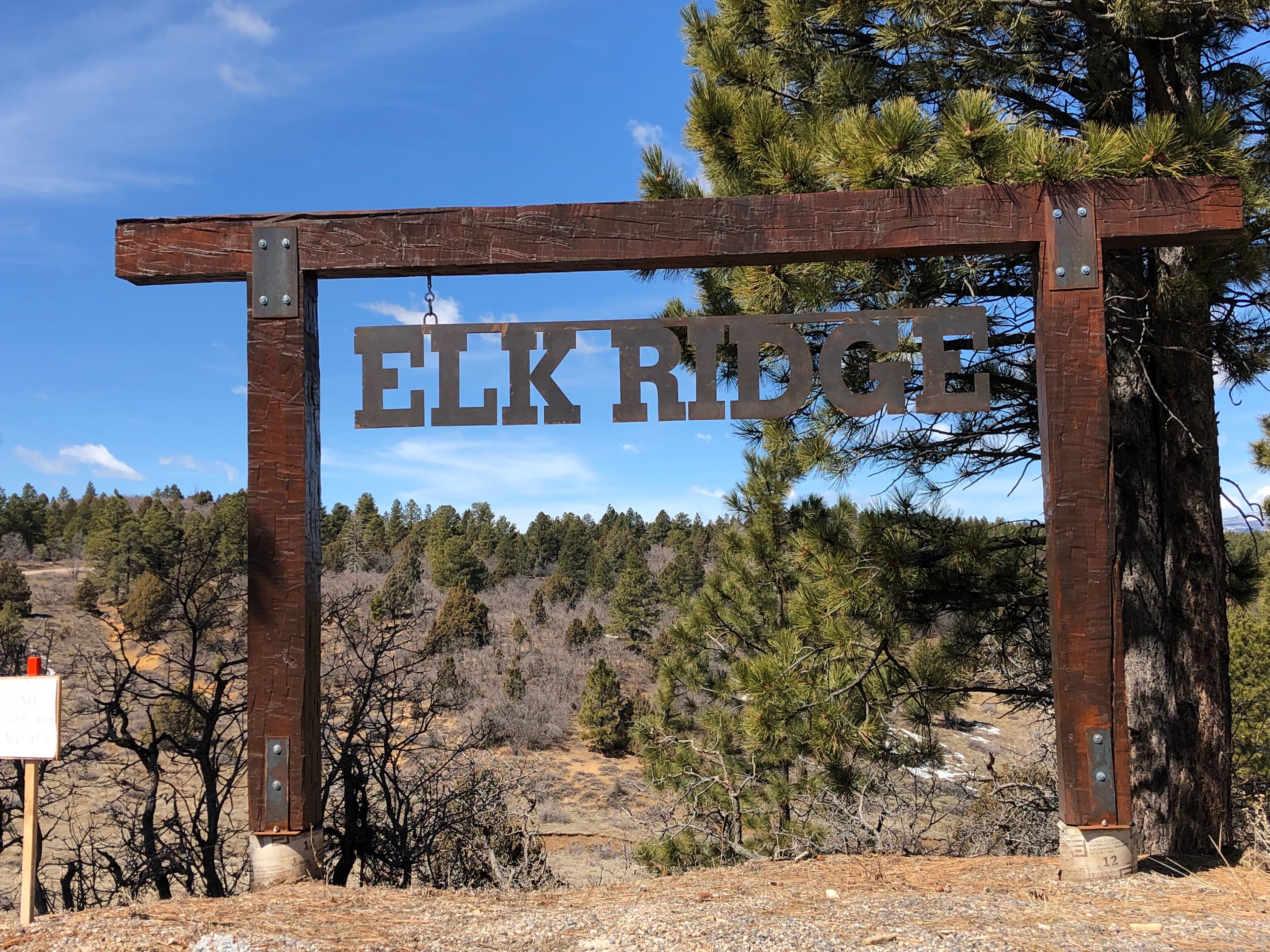 Ein rustikales Holzschild mit dem Schriftzug 'ELK RIDGE' in großen Buchstaben vor einer Baumkulisse und blauem Himmel.