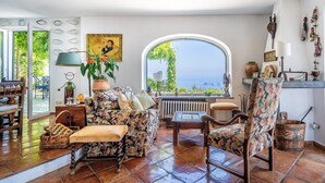 Ein gemütliches Wohnzimmer mit großem Fenster und Meerblick, einer blumigen Couch, einem Holztisch und einem rustikalen Holzstuhl.