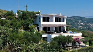 Ein zweistöckiges Haus mit Terrasse, umgeben von viel Grün und mit Blick auf eine hügelige Landschaft.