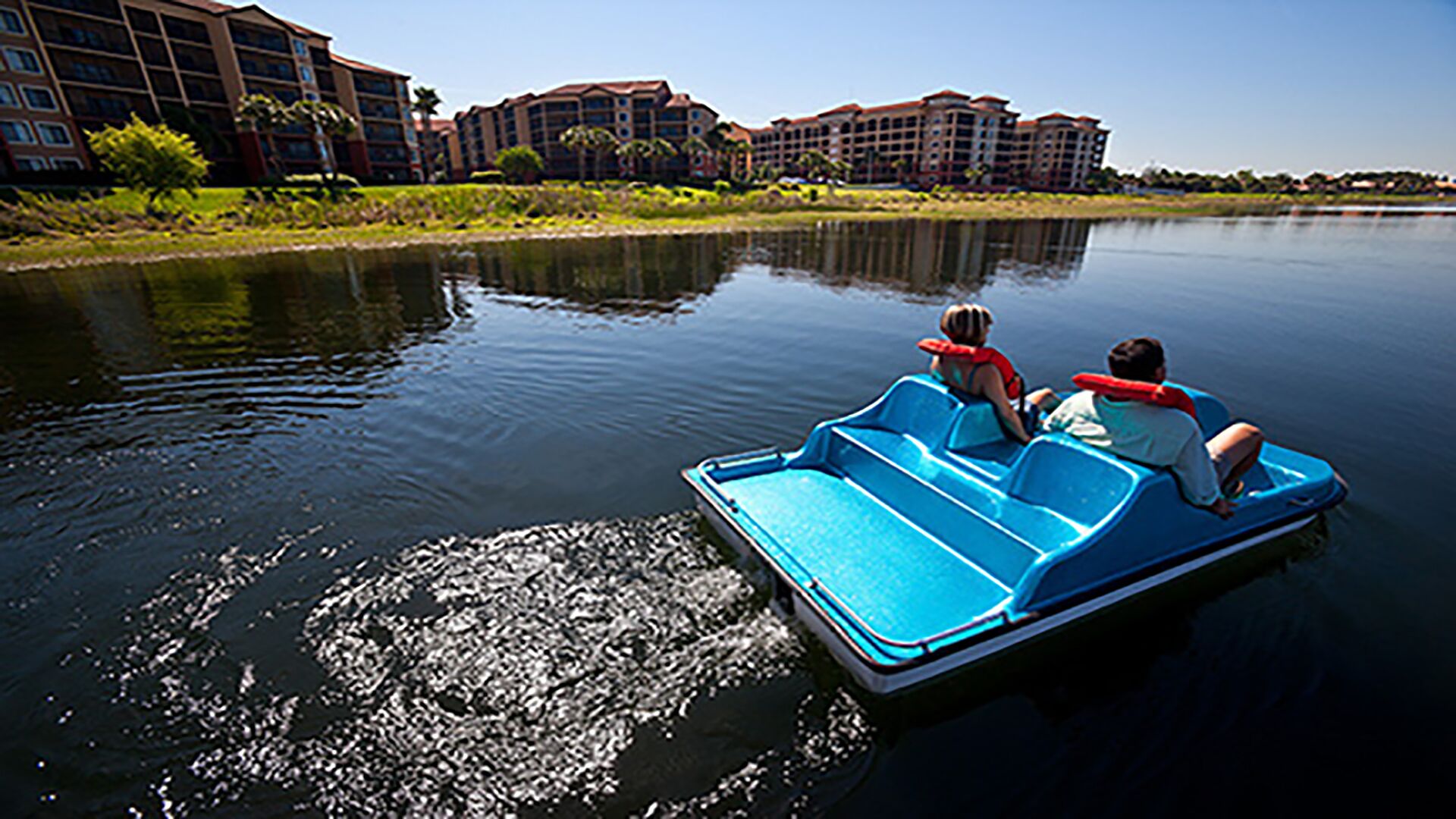 A watercraft with two passengers, a calm lake, and a row of buildings in the background.