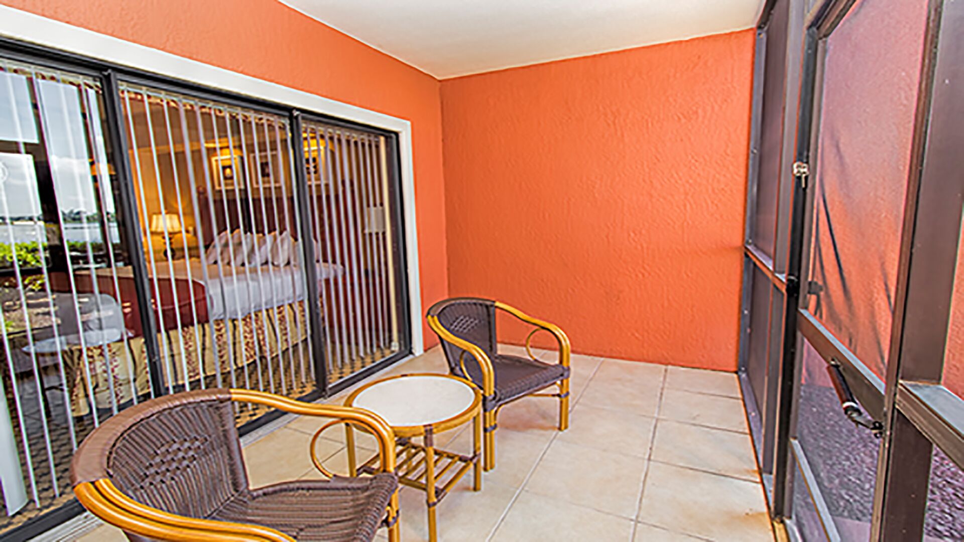 A balcony with wicker chairs and a small table, a sliding glass door, and a view of a room with a bed and curtains.
