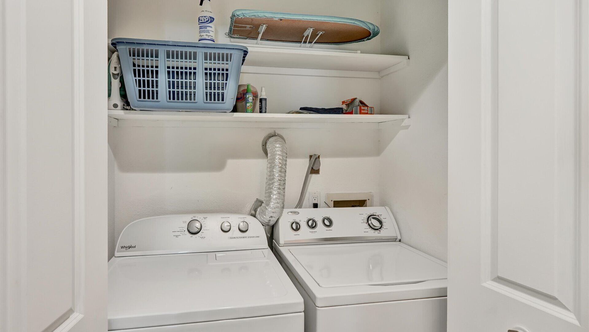 Un cuarto de lavandería con dos lavadoras y secadoras Whirlpool, una cesta de ropa azul y estantes con diversos objetos.
