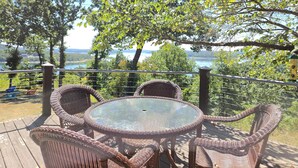 Une terrasse extérieure aménagée avec des meubles en osier et une table en verre, offrant une vue sur un lac et des arbres.