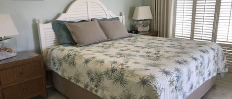 Een slaapkamer met een bed, nachtkastje, lamp en raam met jaloezieën.