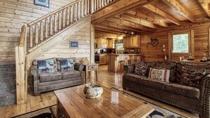 Interior de una cabaña con una escalera de madera, dos sofás con cojines estampados, una zona de cocina y una mesa de comedor.