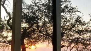 Vista pela janela, emoldurada por madeira, com um pôr do sol sobre um campo verde e árvores.