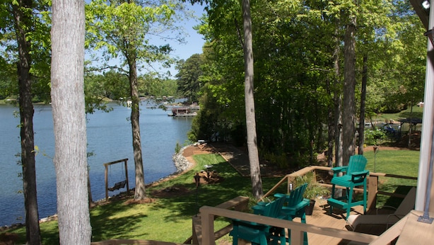 Terraza de madera con sillas verdes con vista a un lago, rodeado de árboles.