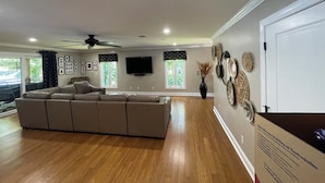 Un salón espacioso con un sofá seccional grande, un ventilador de techo y un televisor de pantalla plana montado en la pared.