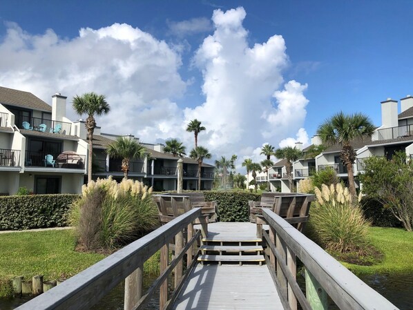 A wooden boardwalk leading to a residential building complex with balconies and palm trees.