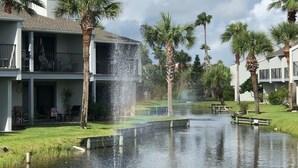 Un quartier résidentiel avec un élément décoratif aquatique au premier plan, entouré de palmiers et de maisons dotées de balcons.