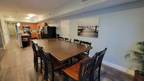 A spacious dining area with a long wooden table and chairs, a kitchen in the background, and a potted plant on the right.