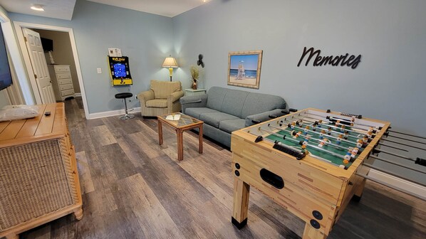 Un salon comprenant une table de baby-foot, un canapé, un fauteuil, une lampe et un téléviseur fixé au mur.