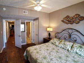 A bedroom with a bed, a nightstand, a lamp, a ceiling fan, and a hallway leading to another room.