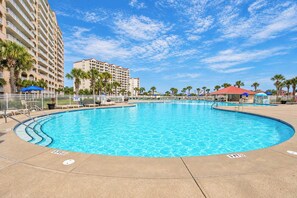 Una gran piscina al aire libre con un solárium circundante, palmeras y un edificio de hotel al fondo.