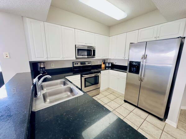 A modern kitchen with white cabinets, stainless steel appliances, a black countertop, and a tiled floor.