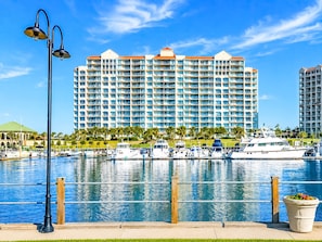 Un hôtel situé au bord de l’eau, doté de nombreux balcons, un port de plaisance avec des bateaux et un ciel d’un bleu éclatant.