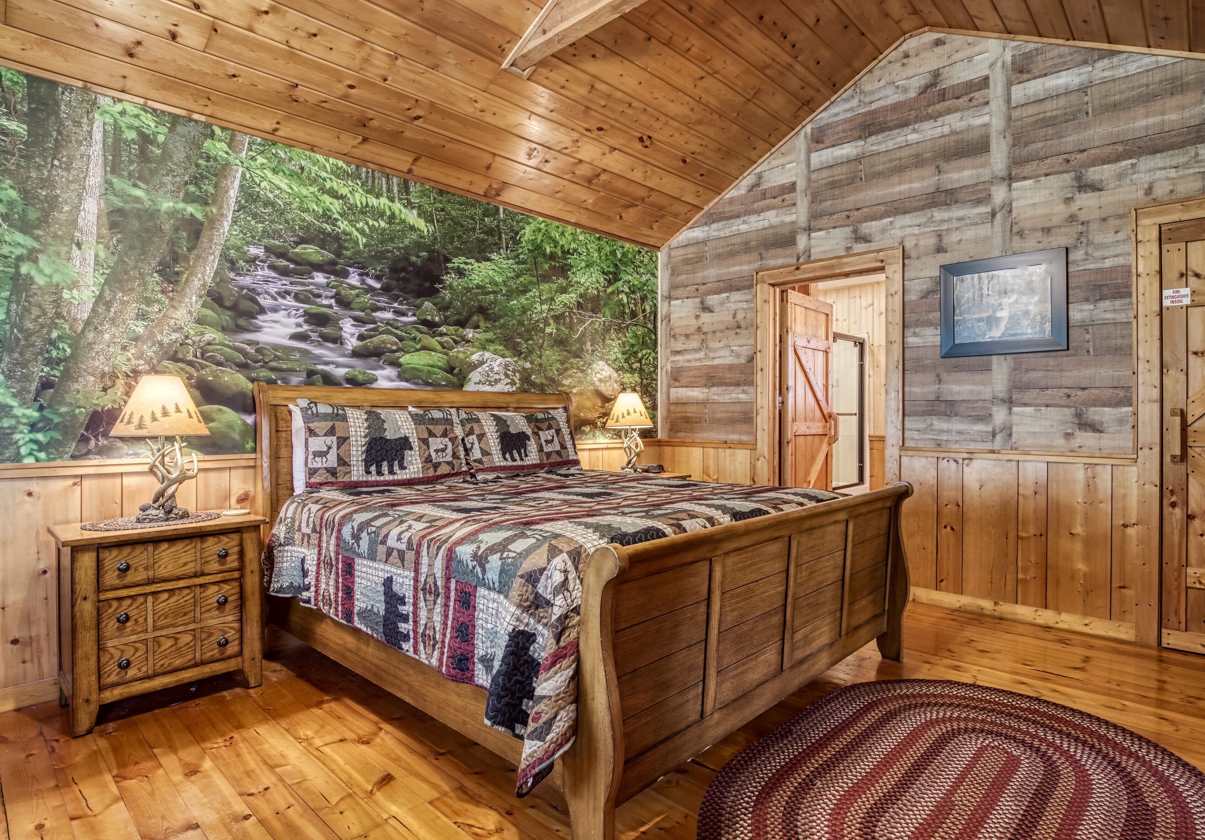 Camera da letto in una baita di legno, con un letto, comodini e un grande murale raffigurante un ruscello in un bosco.